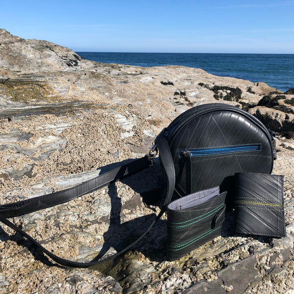 Lifestyle view of a fair trade recycled tyre round shoulder bag with a peacock blue zip. It is on rocks at the beach with the sea in the background and is grouped with two recycled tyre wallets.