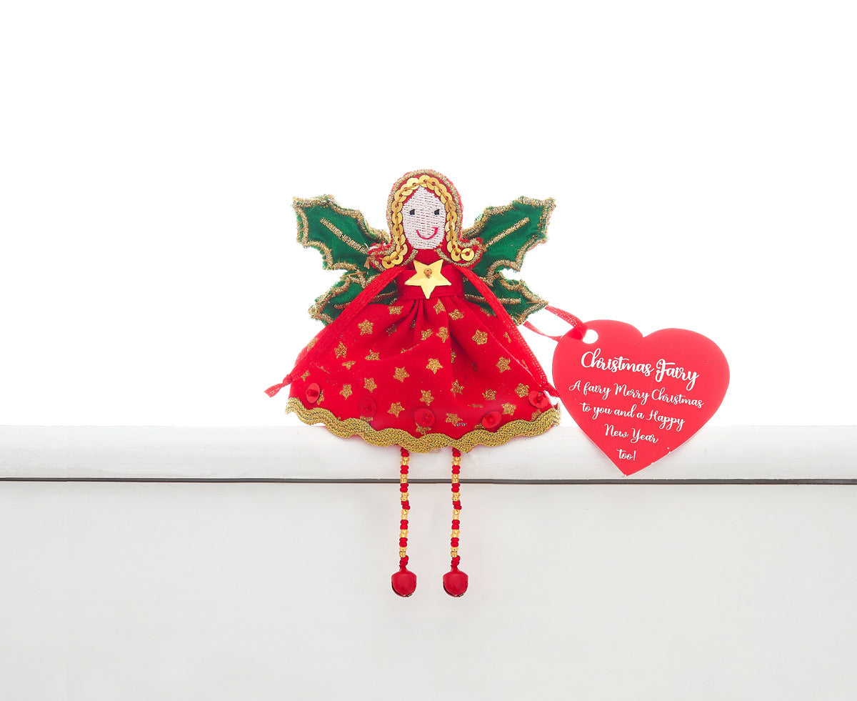 A fair trade handmade Christmas fairy sitting on a white shelf. She is wearing a red dress with  gold stars, red sequins and gold edging and has gold sequin hair and edging on her green holly shaped wings and gold and red beaded legs with red bell feet