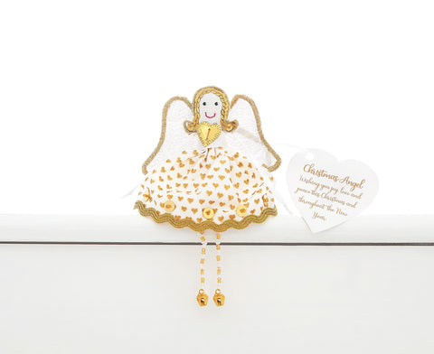 A fair trade handmade Christmas angel sitting on a white shelf. She is wearing a white dress with  gold hearts, sequins and edging and has gold sequin hair and edging on her white wings and gold and white beaded legs with gold bell feet