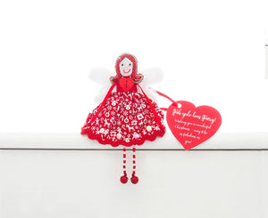A fair trade, handmade, ethical fairy sitting on a white shelf. She is wearing a red dress with white Christmas motifs and a red sequin. She has red sequin hair, white fur wings and red and white beaded legs with red bell feet