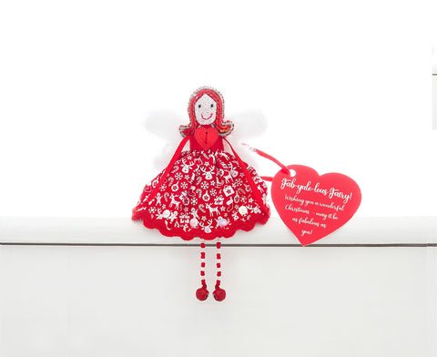 A fair trade, handmade, ethical fairy sitting on a white shelf. She is wearing a red dress with white Christmas motifs and a red sequin. She has red sequin hair, white fur wings and red and white beaded legs with red bell feet