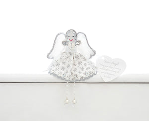 A fair trade handmade Christmas snow angel sitting on a white shelf. She is wearing a white dress with  silver snowflakes, edging and sequins and has silver sequin hair and silver bells on the ends of her silver and white beaded legs