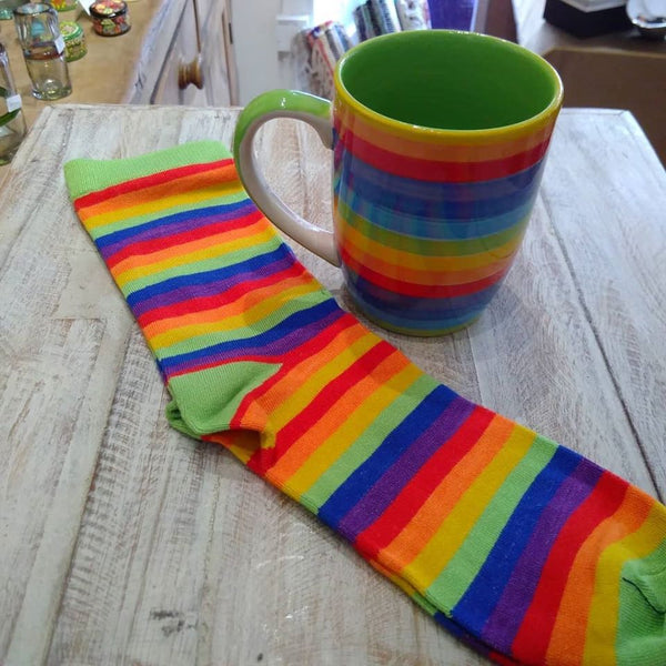 Fair trade bamboo socks with bright rainbow stripes and green heels and toes on a wooden table nest to a fair trade rainbow stripe mug