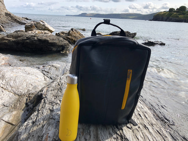 Fair trade, eco friendly, vegan backpack sat on a grey rock with the sea in the background. There is a burnt yellow Chilly's bottle next to the rucksack with the colour matching the yellow of the bag's zip. 