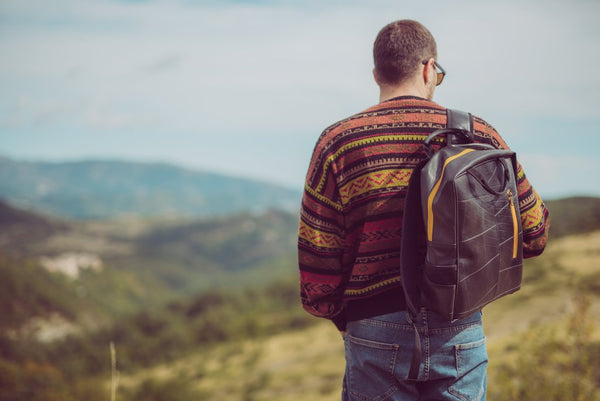 Fair trade, eco friendly, vegan backpack being carried by a male model with white skin and dark hair wearing blue jeans and a patterned jumper. He is standing on a hillside looking out at the view.