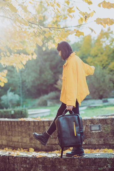 Fair trade, eco friendly, vegan backpack being carried by a female model with white skin and brown hair wearing black leggings and boots and a bright yellow coat. She is walking in a park under yellow autumn leaves.