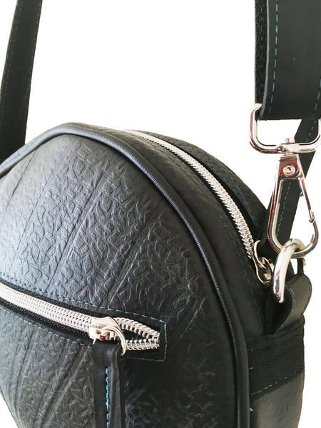 Side view of a fair trade recycled tyre round shoulder bag., showing the front and top zips and the detachable shoulder straps. The bag is black with a texture from the tyre tread and peacock blue stitching around the silver zips and on the straps.