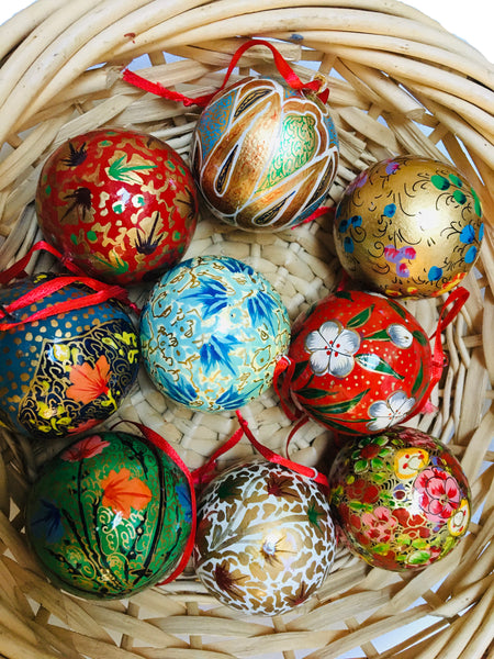 A group of traditional round baubles handpainted with floral patterns in reds, greens, blues and golds. They have a red hanging ribbon and are in a brown wicker basket