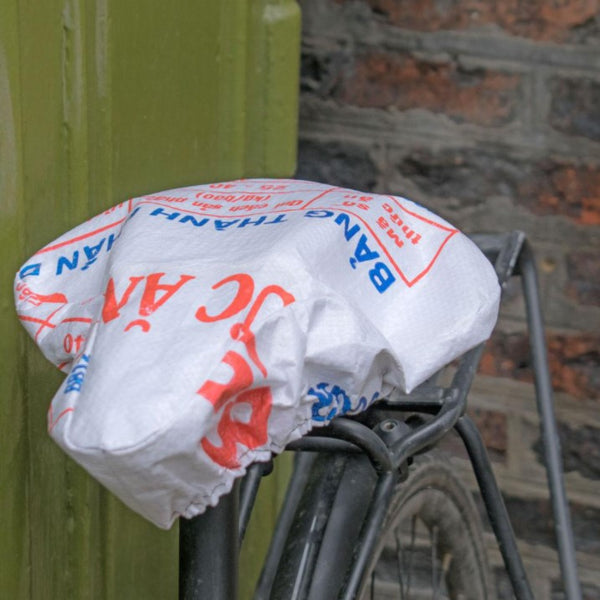 Fair trade and eco friendly recycled cement bag bike seat cover in cream, with red and blue lettering and images from the original cement bag. The cover is shown in place on a black bike leaning against a green gate and a brick wall.