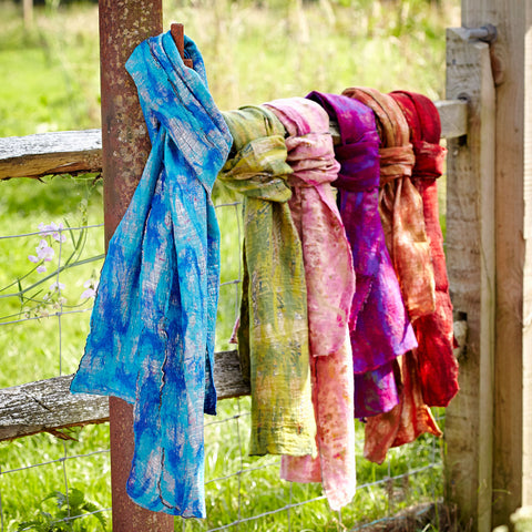 A group of six fair trade silk and felt scarves tied on a fencepost with a meadow in the background. The scarf in the foreground is shades of blue and there are green, pink, purple, orange and red scarves behind it. 