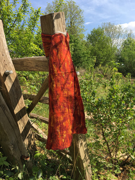 A fair trade silk and felt scarf tied on a wooden fencepost with a summer meadow and trees in the background. The scarf is shades of orange.