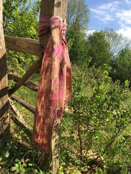 A fair trade silk and felt scarf tied on a wooden fencepost with a summer meadow and trees in the background. The scarf is shades of pink.