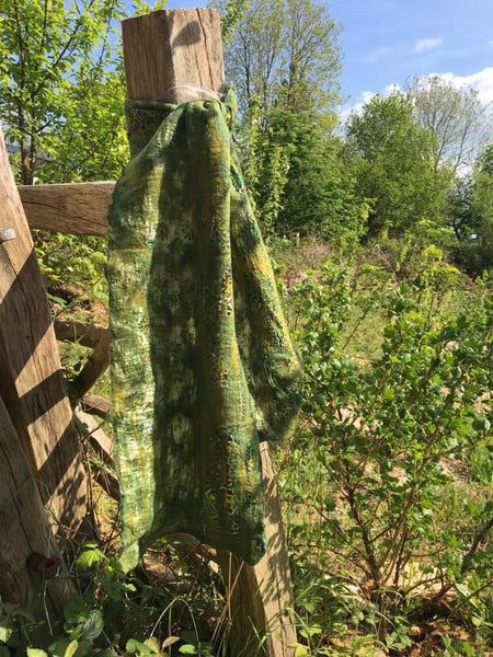 A fair trade silk and felt scarf tied on a wooden fencepost with a summer meadow and trees in the background. The scarf is shades of green.