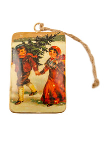 A rectangular tin Christmas decoration showing a vintage scene of a boy and girl carrying a Christmas tree. Main colours are red and green