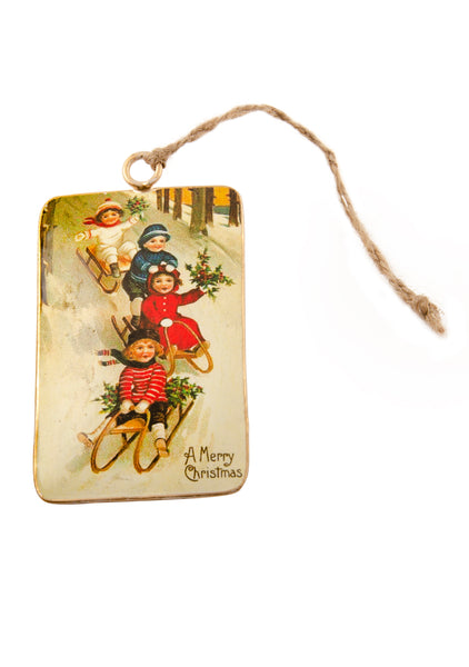 A rectangular tin Christmas decoration showing a vintage scene of four children sledging down a hill. Main colours are red and blue