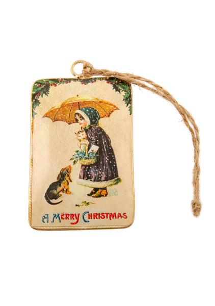 A rectangular tin Christmas decoration showing a vintage scene of a girl with a kitten and a dog under an umbrella in the snow. Main colours are blue and gold